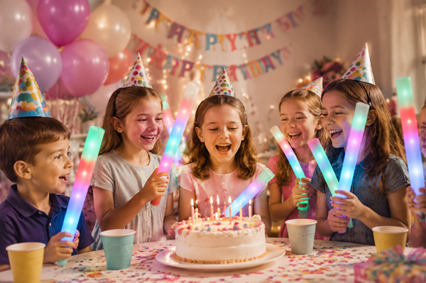 Five children celebrating a birthday and holding glowing light sticks in a decorated party room